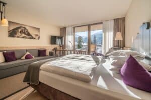 Lehrlingsportal.at Helles Hotelzimmer mit Doppelbett, Sofa, TV, Schreibtisch und Balkon mit Blick auf die Berge. Neutrale Töne mit lila Akzentkissen und Wandkunst eines Berges.