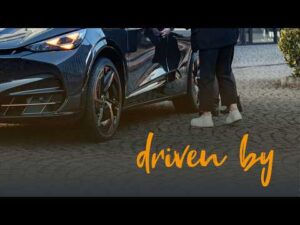 Lehrlingsportal.at Eine Person steht neben einem schwarzen Auto auf einer Kopfsteinpflasterstraße; unten ist in orangefarbener Schrift der Schriftzug „driven by“ zu sehen.