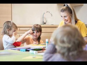 Lehrlingsportal.at Drei kleine Kinder sitzen an einem Tisch mit Bastelmaterial und sind mit einer Bastelarbeit beschäftigt, während eine erwachsene Frau sie in einem Klassenzimmer lächelnd beobachtet.