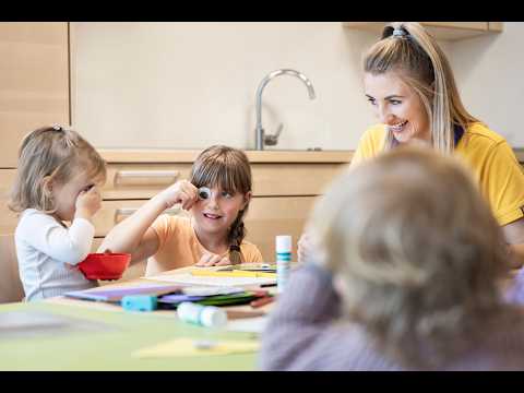 Lehrlingsportal.at Drei kleine Kinder sitzen an einem Tisch mit Bastelmaterial und sind mit einer Bastelarbeit beschäftigt, während eine erwachsene Frau sie in einem Klassenzimmer lächelnd beobachtet.