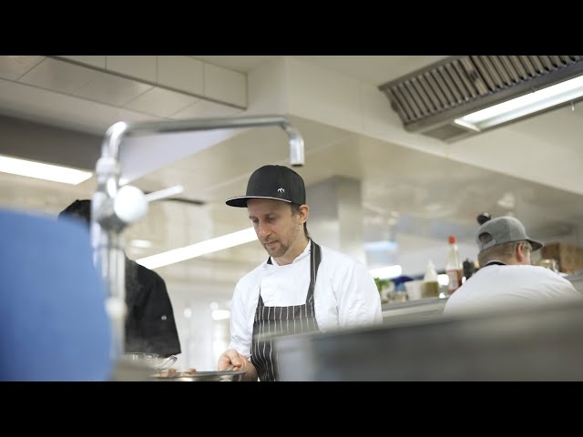 Lehrlingsportal.at Ein Koch in weißer Uniform und schwarzer Mütze arbeitet in einer Großküche und ist mit der Zubereitung von Speisen beschäftigt. Eine weitere Person ist im Hintergrund zu sehen.