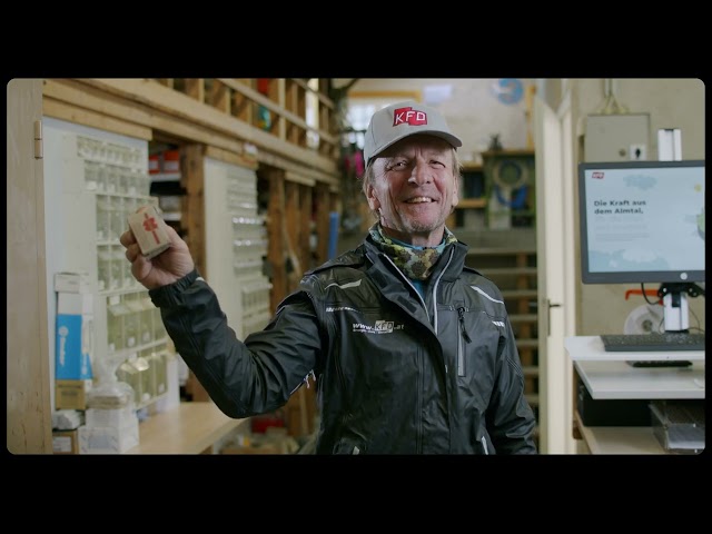 Lehrlingsportal.at Ein älterer Mann mit grauer KFD-Mütze und schwarzer Jacke lächelt, während er in einer Werkstatt oder einem Lagerraum eine kleine Schachtel mit einem roten Kreuz hochhält.