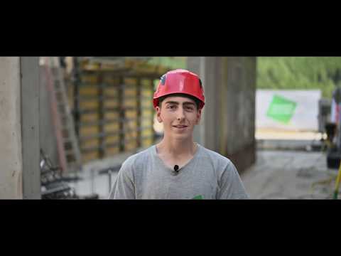 Lehrlingsportal.at Ein junger Mann mit einem roten Schutzhelm und einem grauen Hemd steht auf einer Baustelle und schaut in die Kamera.