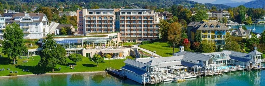 Lehrlingsportal.at Ein großer Hotelkomplex am See mit mehreren Gebäuden, grünen Rasenflächen, Bäumen und Sitzgelegenheiten im Freien, direkt am klaren Wasser und umgeben von einer Stadt und fernen Hügeln.