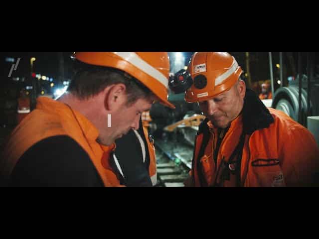 Lehrlingsportal.at Zwei Bauarbeiter in orangefarbener Schutzkleidung stehen nachts neben einem Bahndamm.