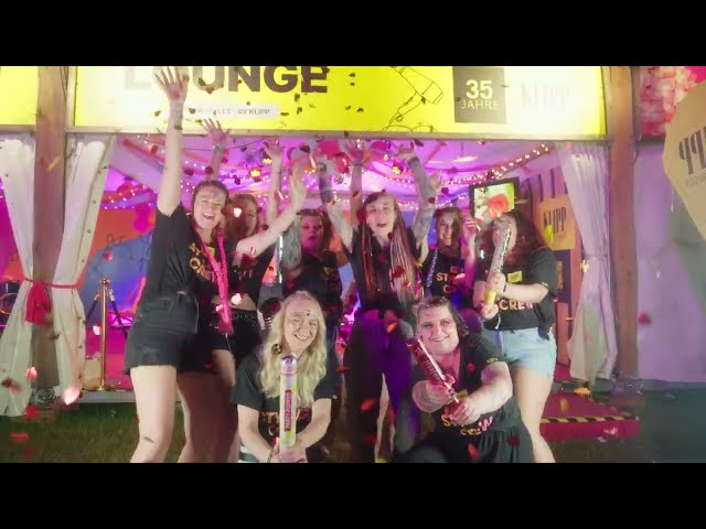 Lehrlingsportal.at Eine Gruppe von Menschen in schwarzen Hemden feiert fröhlich vor einem dekorierten Zelt bei einer Veranstaltung, hält Konfetti-Knaller in der Hand und ist von Konfetti umgeben.