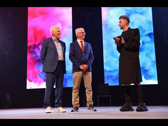 Lehrlingsportal.at Drei Männer stehen auf der Bühne vor bunten Leinwänden. Zwei tragen Anzüge und Turnschuhe, einer einen langen dunklen Mantel. Sie scheinen sich zu unterhalten.