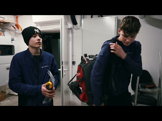 Lehrlingsportal.at Zwei junge Männer in Arbeitsuniformen stehen im Haus; einer hält Werkzeuge, der andere rückt einen Rucksack zurecht.