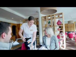 Lehrlingsportal.at Ein Kellner in einem weißen Hemd schenkt einer Frau an einem Restauranttisch Wein ein, während ihr gegenüber eine andere Person sitzt.