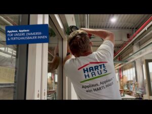 Lehrlingsportal.at Ein Arbeiter in einem "HARTL HAUS"-Hemd baut einen Fensterrahmen in einem modernen Industriegebäude ein. Ein Schild preist Zimmerleute und Fertighausbauer an.