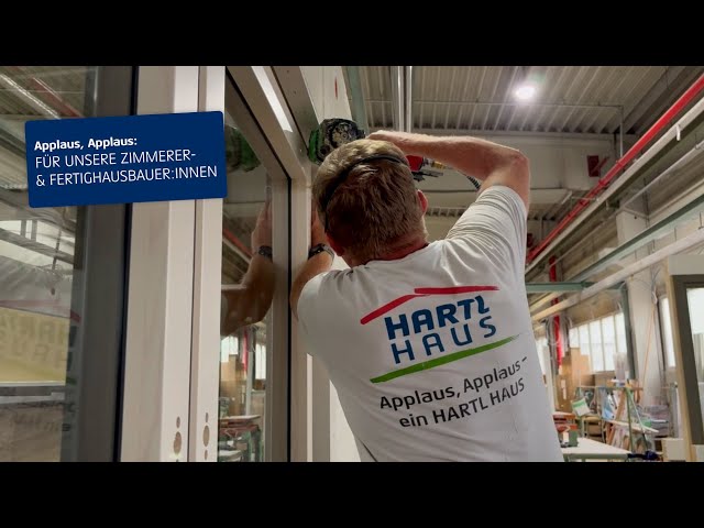 Lehrlingsportal.at Ein Arbeiter in einem "HARTL HAUS"-Hemd baut einen Fensterrahmen in einem modernen Industriegebäude ein. Ein Schild preist Zimmerleute und Fertighausbauer an.