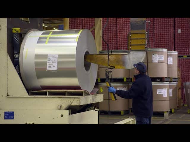 Lehrlingsportal.at Ein Arbeiter in einer Fabrik bedient Maschinen, um eine große Blechrolle zu bewegen, während im Hintergrund weitere Metallrollen zu sehen sind.