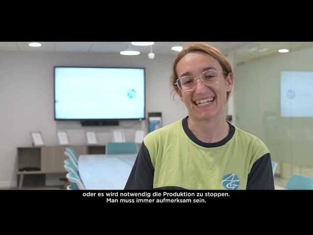 Lehrlingsportal.at Eine Person mit Brille und grünem Hemd steht lächelnd in einem modernen Konferenzraum, mit deutschen Untertiteln am unteren Rand des Bildes.