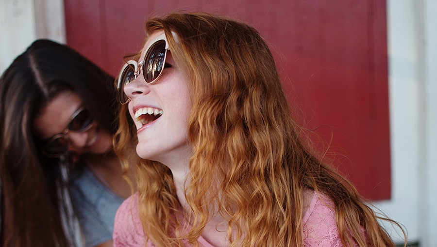 Lehrlingsportal.at Zwei Menschen mit langen Haaren und Sonnenbrillen lachen. Die Person im Vordergrund hat rote Haare und trägt ein rosa Oberteil.