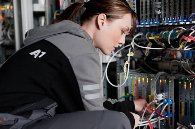 Lehrlingsportal.at Eine Person in einem grauen Kapuzenpullover arbeitet an der Verkabelung in einem Serverraum, umgeben von verschiedenen Kabeln und Geräten.