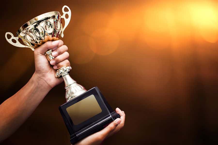 Lehrlingsportal.at Eine Hand hält eine goldene Trophäe mit einer leeren Plakette vor einem warmen, leuchtenden Hintergrund hoch.