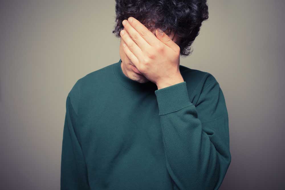 Lehrlingsportal.at Eine Person in einem grünen Pullover verdeckt ihr Gesicht mit einer Hand und steht vor einem einfarbigen Hintergrund.