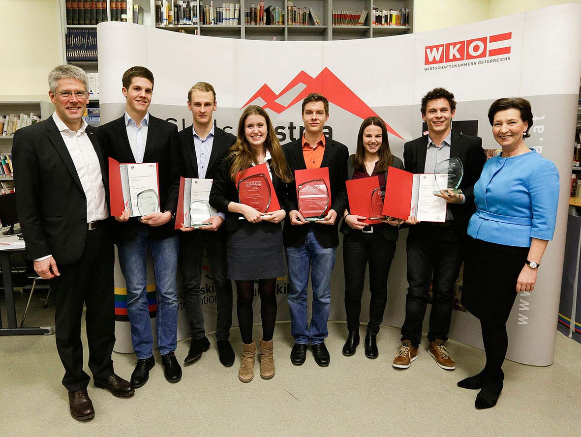Lehrlingsportal.at Sieben Personen stehen in einer Reihe und halten Auszeichnungen in den Händen, flankiert von zwei weiteren Personen. Sie stehen vor einem Hintergrund mit Logos und roten dreieckigen Mustern.