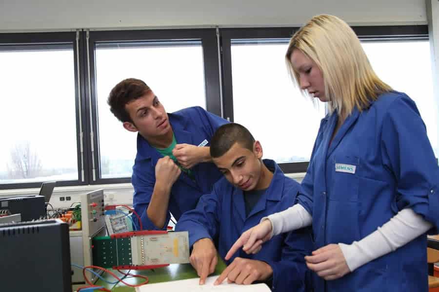 Lehrlingsportal.at Drei Personen in blauen Laborkitteln arbeiten gemeinsam an einem elektronischen Projekt, diskutieren und zeigen auf ein Dokument.