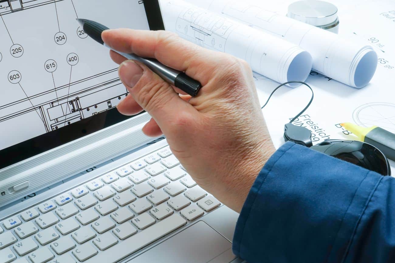 Lehrlingsportal.at Eine Person mit einem Stift zeigt auf Baupläne auf einem Laptop-Bildschirm. Im Hintergrund sind aufgerollte Baupläne und eine Tastatur zu sehen.