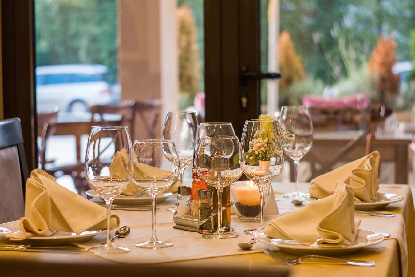 Lehrlingsportal.at Ein hübsch gedeckter Esstisch mit gefalteten Servietten, Weingläsern, einer Kerze und einem kleinen Blumengesteck in einem Restaurant mit Blick ins Freie.