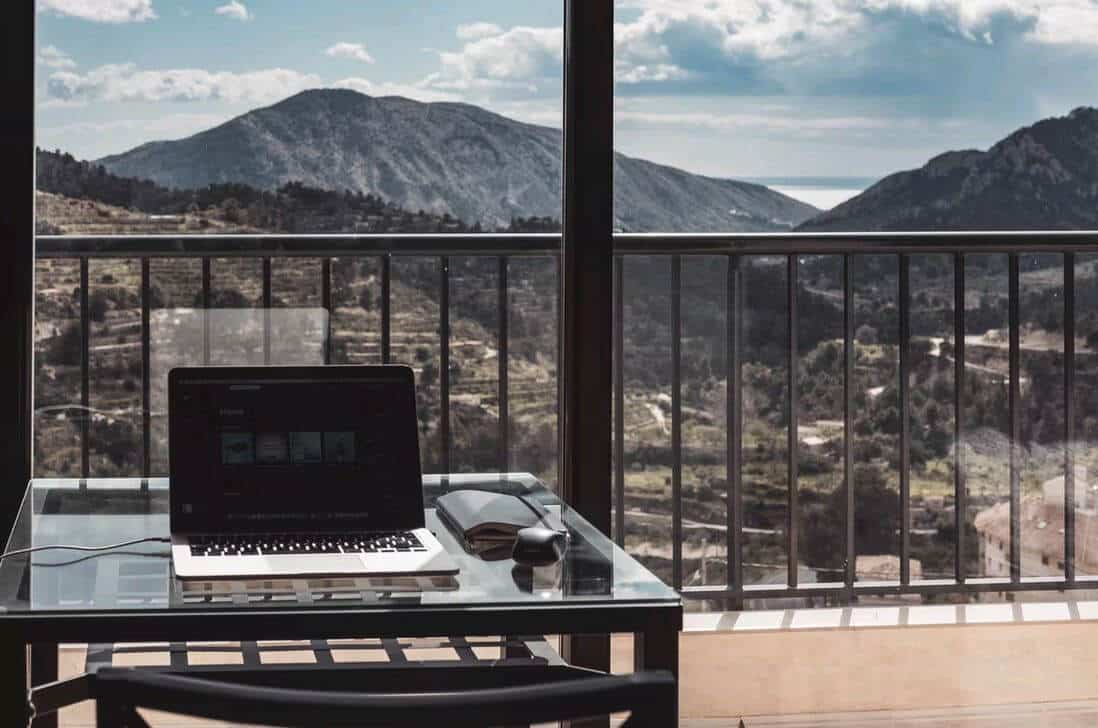 Lehrlingsportal.at Ein Laptop mit Maus und Notizbuch auf einem Glastisch mit Blick auf einen Balkon mit Bergen und einem bewölkten Himmel.