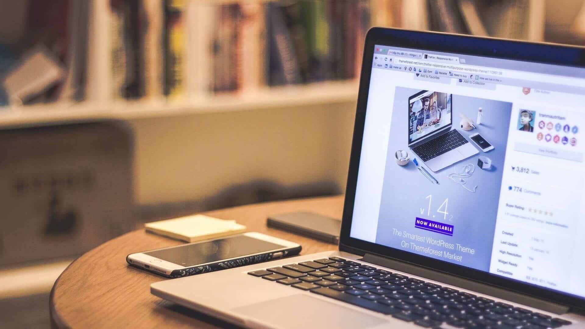 Lehrlingsportal.at Offener Laptop auf einem Holztisch mit Smartphone, Notizbuch und Buch, im Hintergrund Bücherregale.