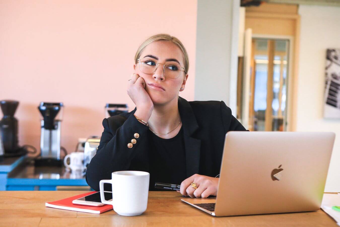 Lehrlingsportal.at Person im Blazer sitzt an einem Tisch, stützt das Kinn auf die Hand, vor ihr liegen ein Laptop, eine Kaffeetasse und Notizbücher. Wirkt nachdenklich in einer Büroumgebung.