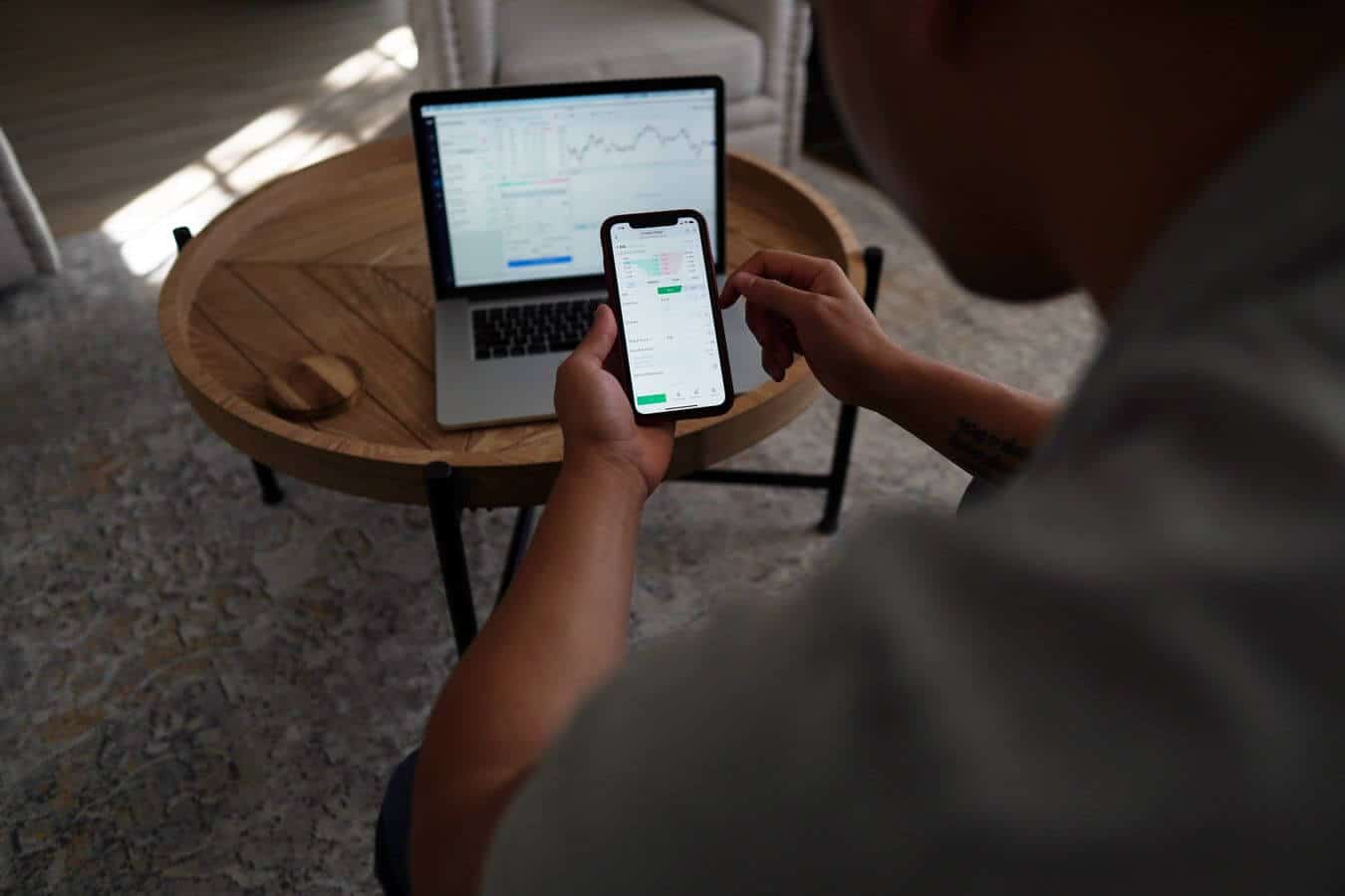 Lehrlingsportal.at Person sitzt an einem runden Holztisch und betrachtet auf einem Smartphone und Laptop Grafiken und Diagramme.