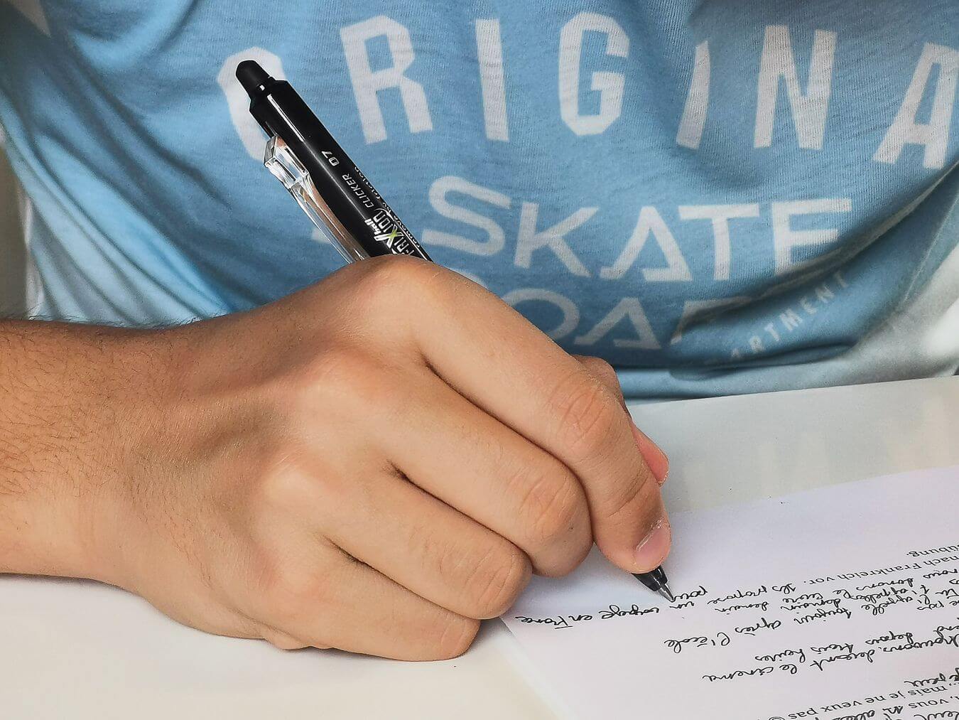 Lehrlingsportal.at Eine Person schreibt mit einem schwarzen Stift auf einem Papier und trägt ein hellblaues Hemd mit weißer Schrift.