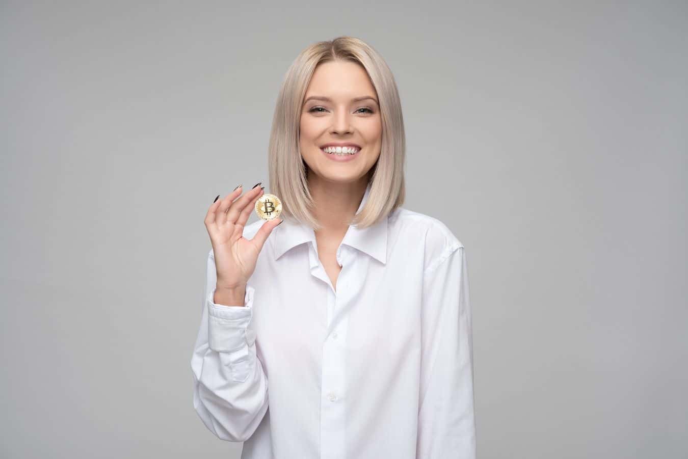 Lehrlingsportal.at Eine Person mit blonden Haaren und weißem Hemd hält eine Bitcoin-Münze und lächelt vor einem grauen Hintergrund.