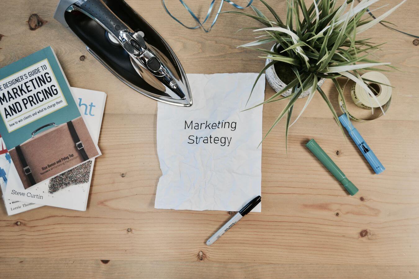 Lehrlingsportal.at Ein zerknülltes Papier mit dem Text „Marketingstrategie“ auf einem Schreibtisch, umgeben von einem Bügeleisen, einer Pflanze, einem Buch, einem Stift, Klebeband und einem Textmarker.