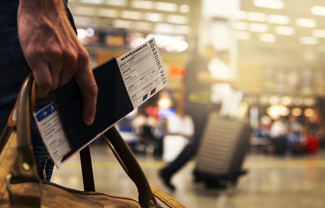 Lehrlingsportal.at Person mit Reisepass und Bordkarte an einem Flughafen, im Hintergrund sind unscharfe Reisende und ein Koffer zu sehen.