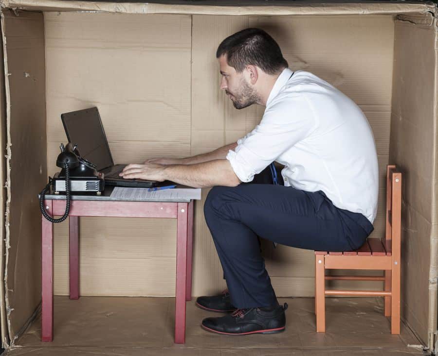 Lehrlingsportal.at Ein Mann sitzt auf einem kleinen Stuhl an einem kompakten Schreibtisch in einem Karton und arbeitet an einem Laptop.