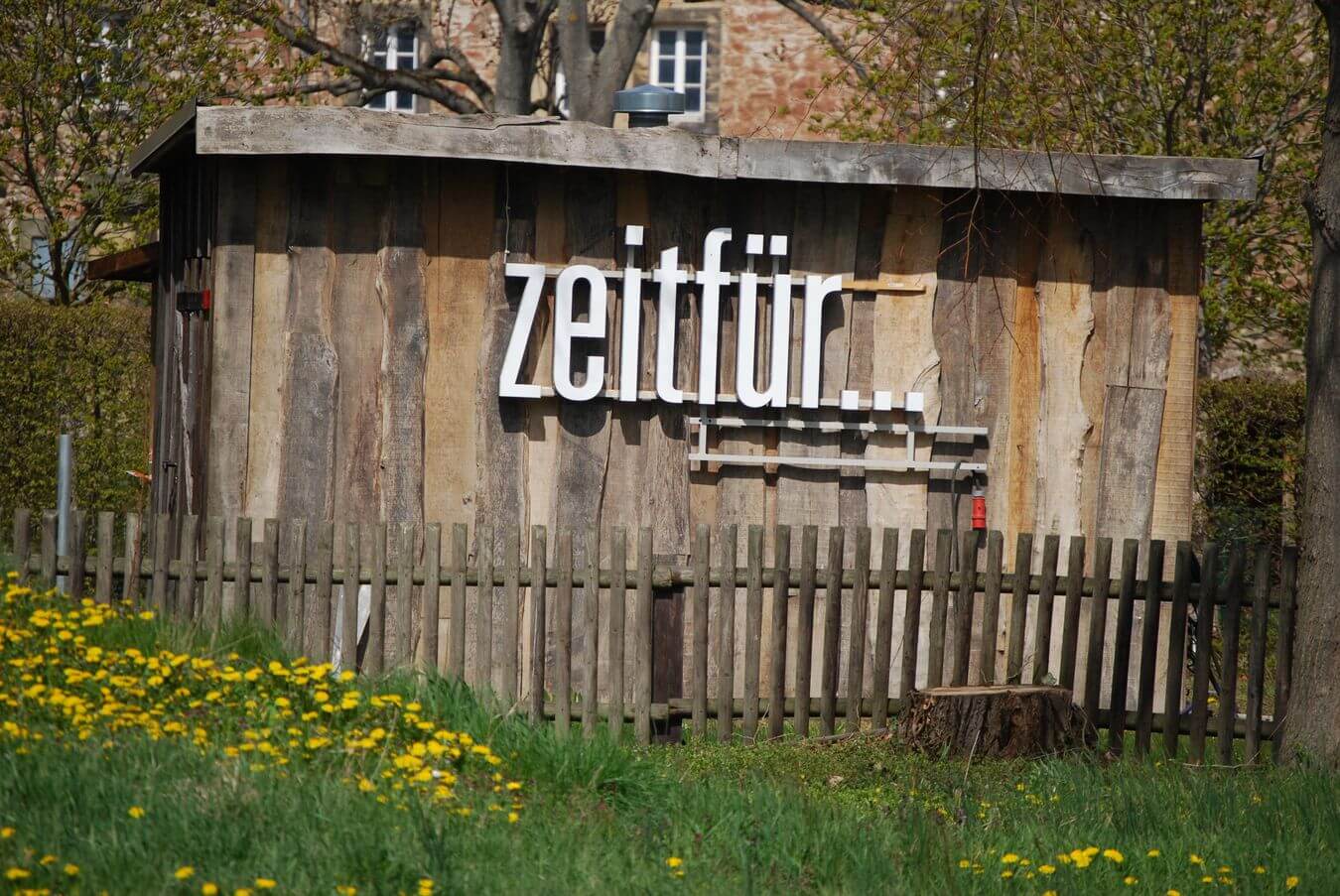 Lehrlingsportal.at Ein rustikaler Holzschuppen mit der Aufschrift „zeitfür…“ an der Wand, umgeben von einem Holzzaun und einem Feld mit gelben Blumen.
