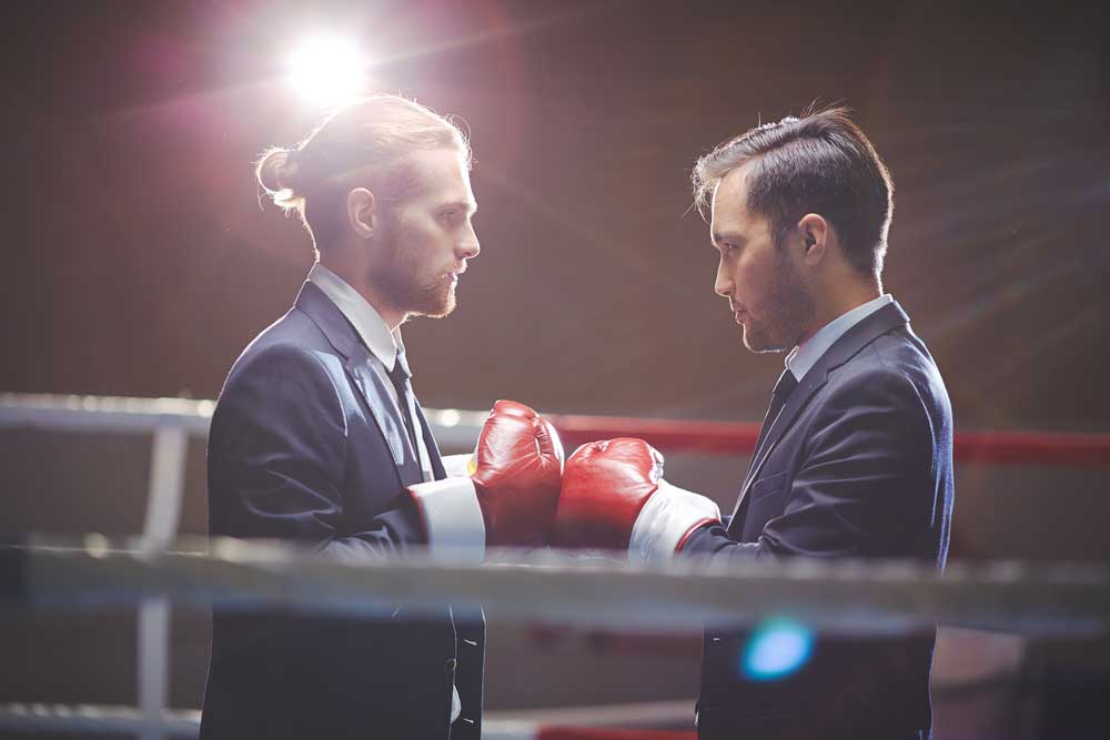 Lehrlingsportal.at In einem Boxring stehen sich zwei Männer in Anzügen gegenüber. Sie tragen rote Handschuhe und hinter ihnen leuchtet ein helles Licht.