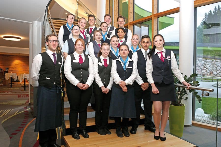 Lehrlingsportal.at Eine Gruppe uniformierter Mitarbeiter steht auf einer Treppe und posiert für ein Gruppenfoto. Sie lächeln alle, und im Hintergrund ist ein modernes Interieur mit großen Fenstern zu sehen.