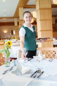 Lehrlingsportal.at Ein Kellner in einer grünen Weste hält ein Tablett und arrangiert die Tische in einem gut beleuchteten Restaurantbereich. Sonnenblumen schmücken die Tische.