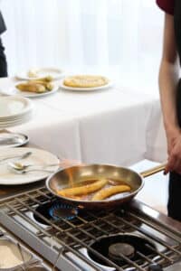Lehrlingsportal.at Eine Person kocht Bananen in einer Pfanne auf einem Herd, die Flammen sind sichtbar. In der Nähe ist ein Tisch mit Tellern und Essen gedeckt.