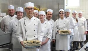 Lehrlingsportal.at Eine Gruppe von Köchen in weißen Uniformen und Hüten steht in einer Küche und hält Tabletts mit Meeresfrüchten mit der Aufschrift „Fine De Claire“.