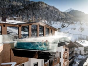 Lehrlingsportal.at Ein modernes Gebäude mit einem über den Rand hinausragenden Pool mit Glasboden, umgeben von schneebedeckten Bergen und Tannen unter einem klaren Himmel. Zwei Personen sind im Pool.