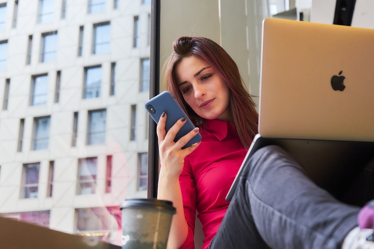 Lehrlingsportal.at Frau in rotem Hemd sitzt mit Smartphone und Laptop neben einer Kaffeetasse. Durch das große Fenster hinter ihr ist die Fassade eines modernen Gebäudes zu sehen.