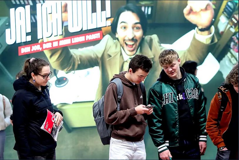 Lehrlingsportal.at Drei junge Menschen stehen vor einer großen Anzeige mit einem enthusiastisch posierenden Mann und dem deutschen Text "JA! ICH WILL!" in fetten Buchstaben.