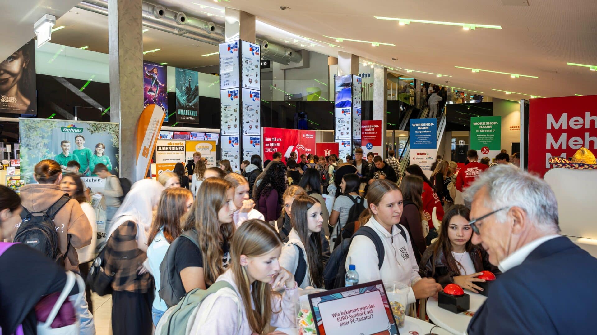 Lehrlingsportal.at Eine große Gruppe von Menschen, vor allem junge Frauen, versammelt sich in einem hell beleuchteten Veranstaltungsraum mit Informationsständen und Plakaten.