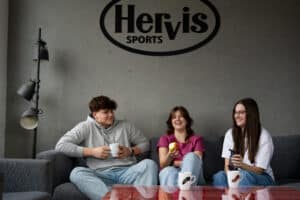 Lehrlingsportal.at Drei Personen sitzen mit Tassen auf einem grauen Sofa, eine davon hält einen Apfel, unter einem „Hervis Sports“-Schild an einer grauen Wand.