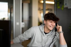Lehrlingsportal.at Ein junger Mann in einem grauen Kapuzenpullover lächelt, während er in einem Büro mit einem schnurgebundenen Telefon telefoniert.