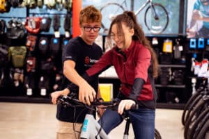 Lehrlingsportal.at Zwei Personen stehen in einem Fahrradladen. Eine sitzt auf einem Fahrrad, die andere steht daneben und hilft beim Einstellen des Lenkers. Im Hintergrund sind Regale mit Fahrradzubehör zu sehen.