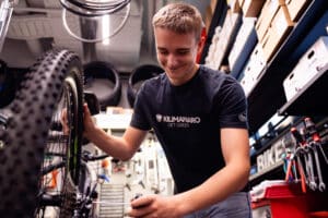 Lehrlingsportal.at Ein junger Mann stellt in einer Werkstatt ein Mountainbike-Rad ein, umgeben von Werkzeugen und Regalen mit Kisten.