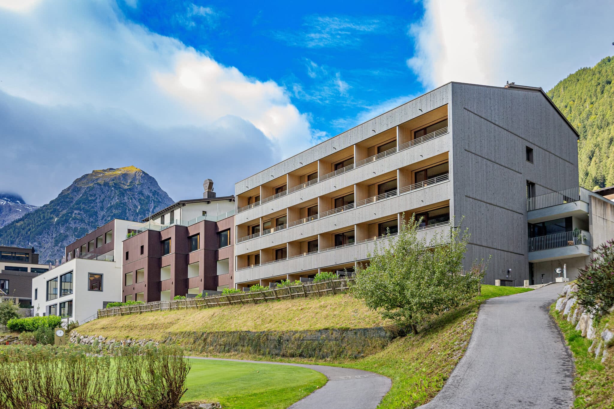 Lehrlingsportal.at Modernes mehrstöckiges Hotelgebäude mit Balkonen, an einem Hang gelegen, mit gepflastertem Weg, viel Grün und Bergen im Hintergrund unter einem teilweise bewölkten Himmel.