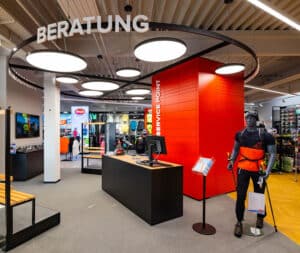 Lehrlingsportal.at Im Inneren eines Einzelhandelsgeschäfts gibt es einen Beratungsschalter mit der Aufschrift „Beratung“, eine rote Service-Wand und eine Schaufensterpuppe in Outdoor-Ausrüstung, die Kletterausrüstung hält.
